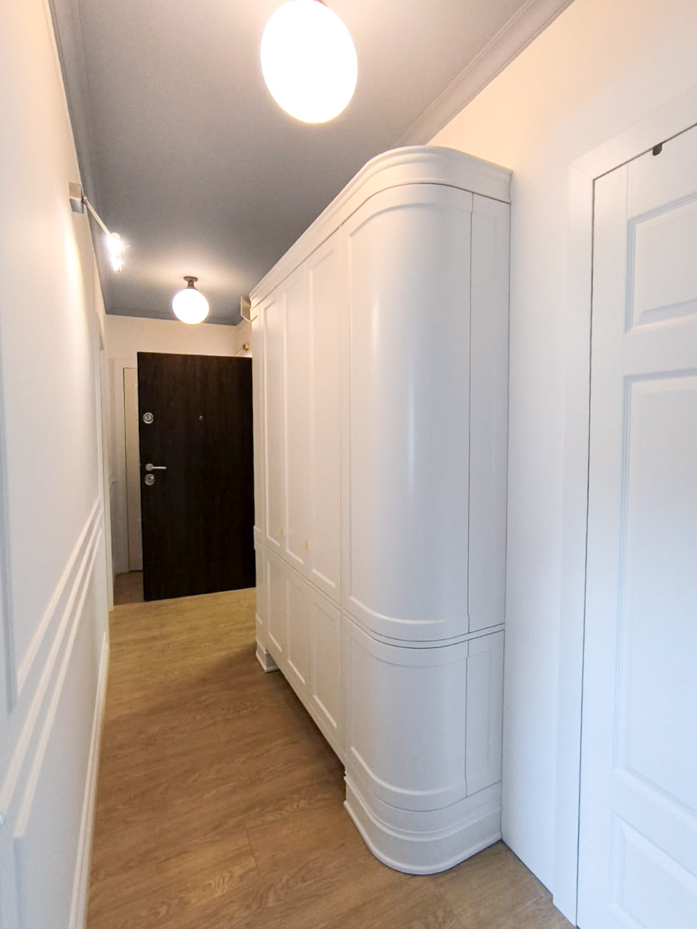Biała szafa z zaokrąglonymi bokami w wąskim korytarzu z drewnianą podłogą. Widoczne drzwi wejściowe i oświetlenie sufitowe. Minimalistyczny design.