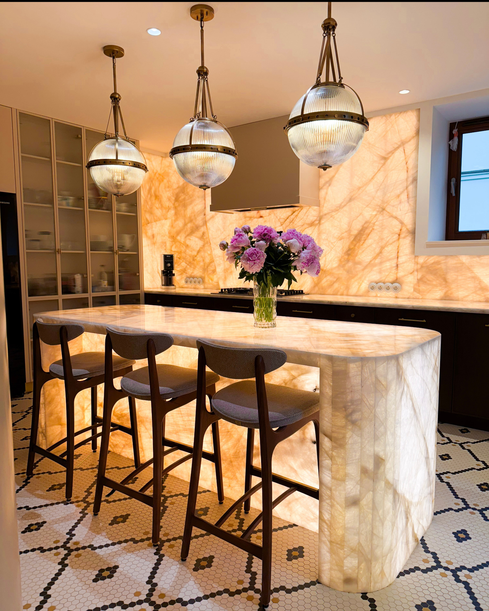 Nowoczesna kuchnia z podświetlanym blatem z onyksu, trzema wiszącymi lampami w stylu retro i mozaikową podłogą. Na blacie wazon z różowymi piwoniami.