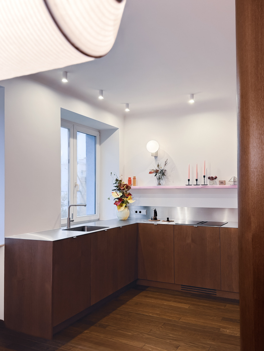 Nowoczesna kuchnia z drewnianymi frontami, stalowym blatem i oknem. Minimalistyczny design z akcentami kolorystycznymi na półce i dekoracjami. Oświetlenie punktowe.