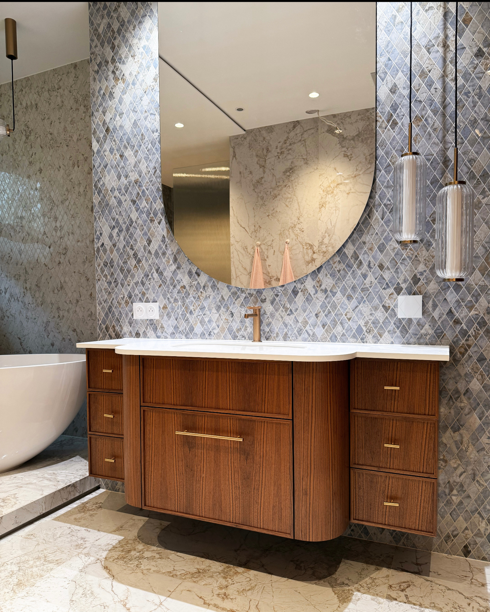 Nowoczesna łazienka z drewnianą szafką pod umywalką, owalnym lustrem i mozaiką na ścianie. Widoczne wiszące lampy i fragment wanny. Całość w ciepłej tonacji.