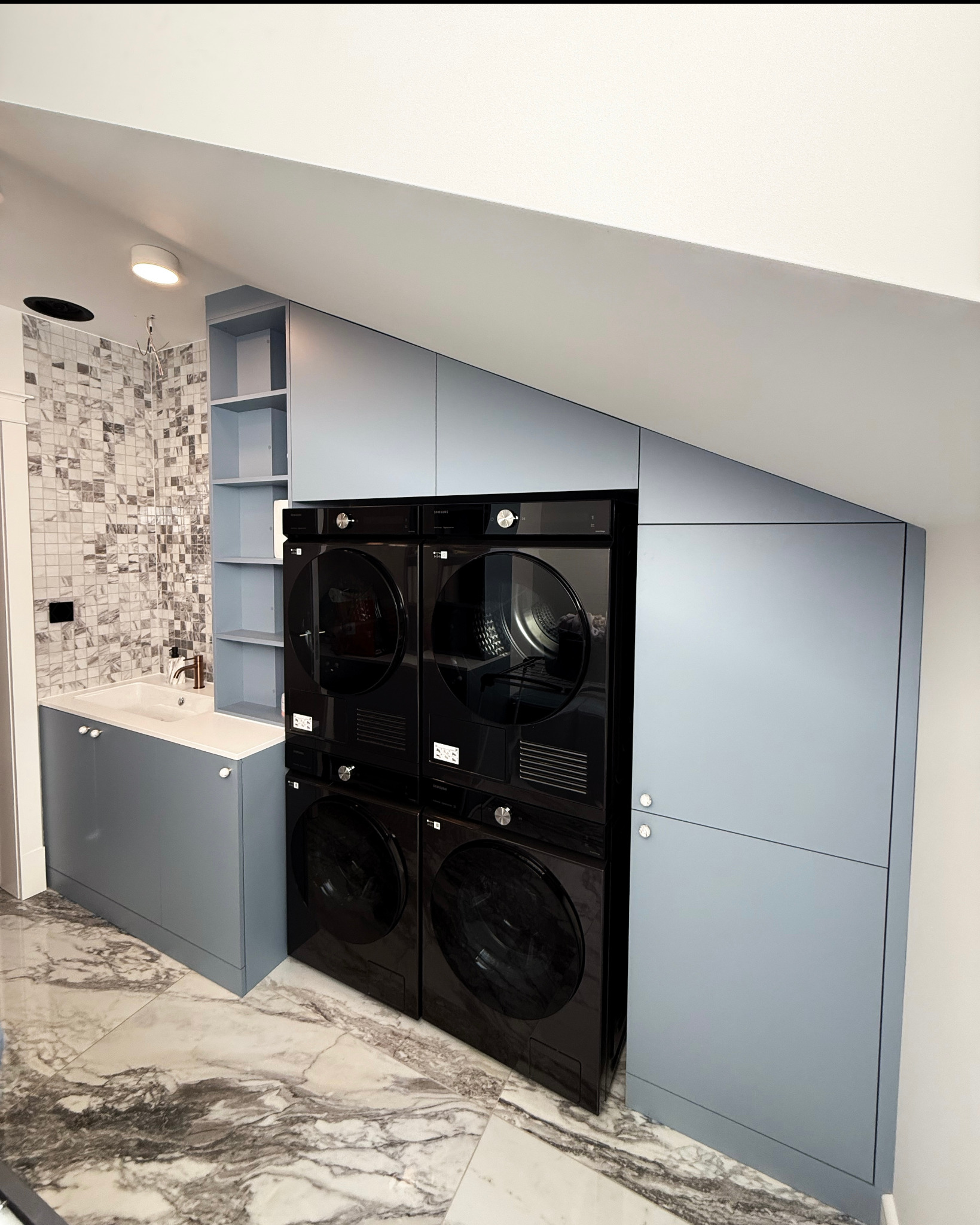 Łazienka na poddaszu z zabudową meblową w odcieniach szarości, marmurową podłogą i mozaiką na ścianie. Uwagę przyciąga pionowy zestaw pralki i suszarki.