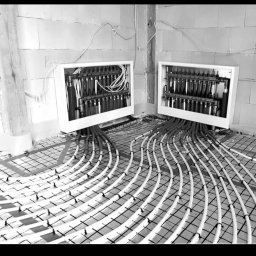 DIAMENT TOMASZ GUZEWSKI - Czarno-białe zdjęcie instalacji ogrzewania podłogowego z rozdzielaczami w surowym wnętrzu. Widoczne rury ułożone w pętle na siatce montażowej, przygotowane do zalania wylewką.