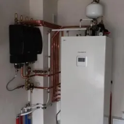 Zainstalowana biała pompa ciepła LG Therma V z podłączonymi miedzianymi rurami w pomieszczeniu technicznym. Widoczne czarne moduły i biały zbiornik wyrównawczy.