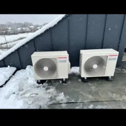 Dwa białe klimatyzatory Toshiba Inverter stoją na zewnątrz, na dachu pokrytym śniegiem. Widoczne drzewa w tle. Zimowa instalacja klimatyzacji.