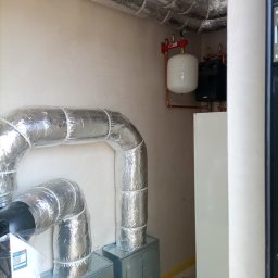 DIAMENT TOMASZ GUZEWSKI - Izolowane rury wentylacyjne i elementy instalacji grzewczej w pomieszczeniu technicznym, widoczne zbiornik wyrównawczy i fragment kotła. Szare ściany, surowy stan wykończenia.