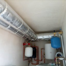 DIAMENT TOMASZ GUZEWSKI - Złożona instalacja grzewcza z rurami w izolacji aluminiowej, zbiornikami wyrównawczymi (biały i niebieski) oraz kotłem, zamontowana w pomieszczeniu technicznym.