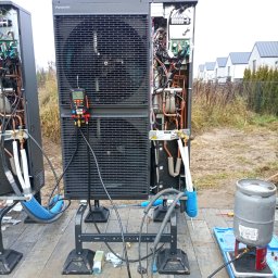 DIAMENT TOMASZ GUZEWSKI - Dwie pompy ciepła Panasonic na zewnątrz, jedna z otwartą obudową, podłączone narzędzia pomiarowe i butla z gazem. Widoczne domy w tle.