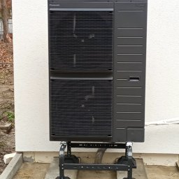 DIAMENT TOMASZ GUZEWSKI - Czarna pompa ciepła Panasonic zamontowana na zewnątrz budynku na specjalnej konstrukcji wsporczej. Widoczna ściana budynku z jasnym tynkiem i fragment ogrodu.