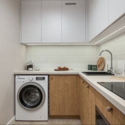 Karol Lis - Nowoczesna kuchnia z pralką w zabudowie, drewniane fronty szafek, białe blaty i górne szafki, oświetlenie LED pod szafkami, minimalistyczny design, płytki z subtelnym wzorem.