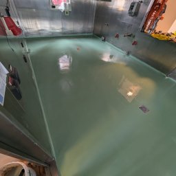 Karol Lis - Świeżo pomalowana, gładka, zielona podłoga w pomieszczeniu o metalowych ścianach. Widoczne narzędzia i wiadro z farbą. Oświetlenie odbija się od mokrej powierzchni.