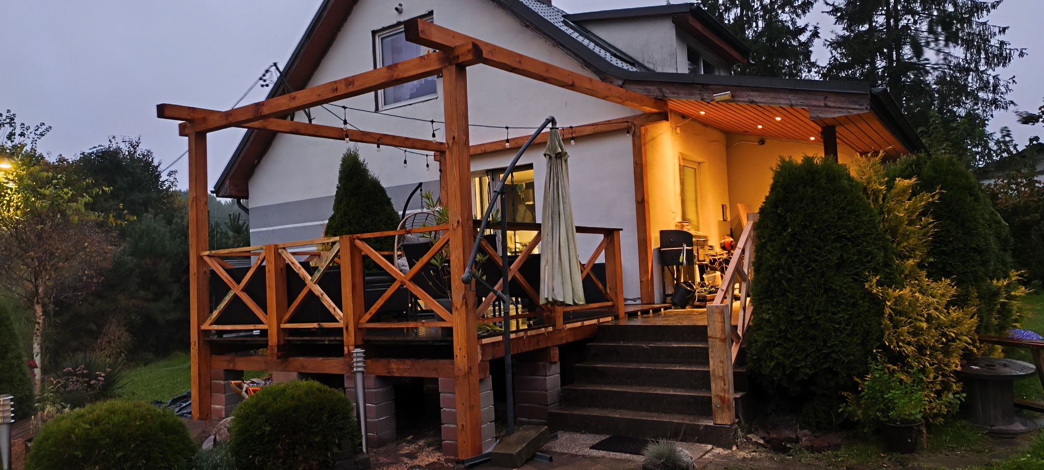 Drewniany taras z balustradą przy domu jednorodzinnym, oświetlony girlandami żarówek. Widoczne schody wejściowe i elementy dekoracyjne, takie jak parasol i rośliny.