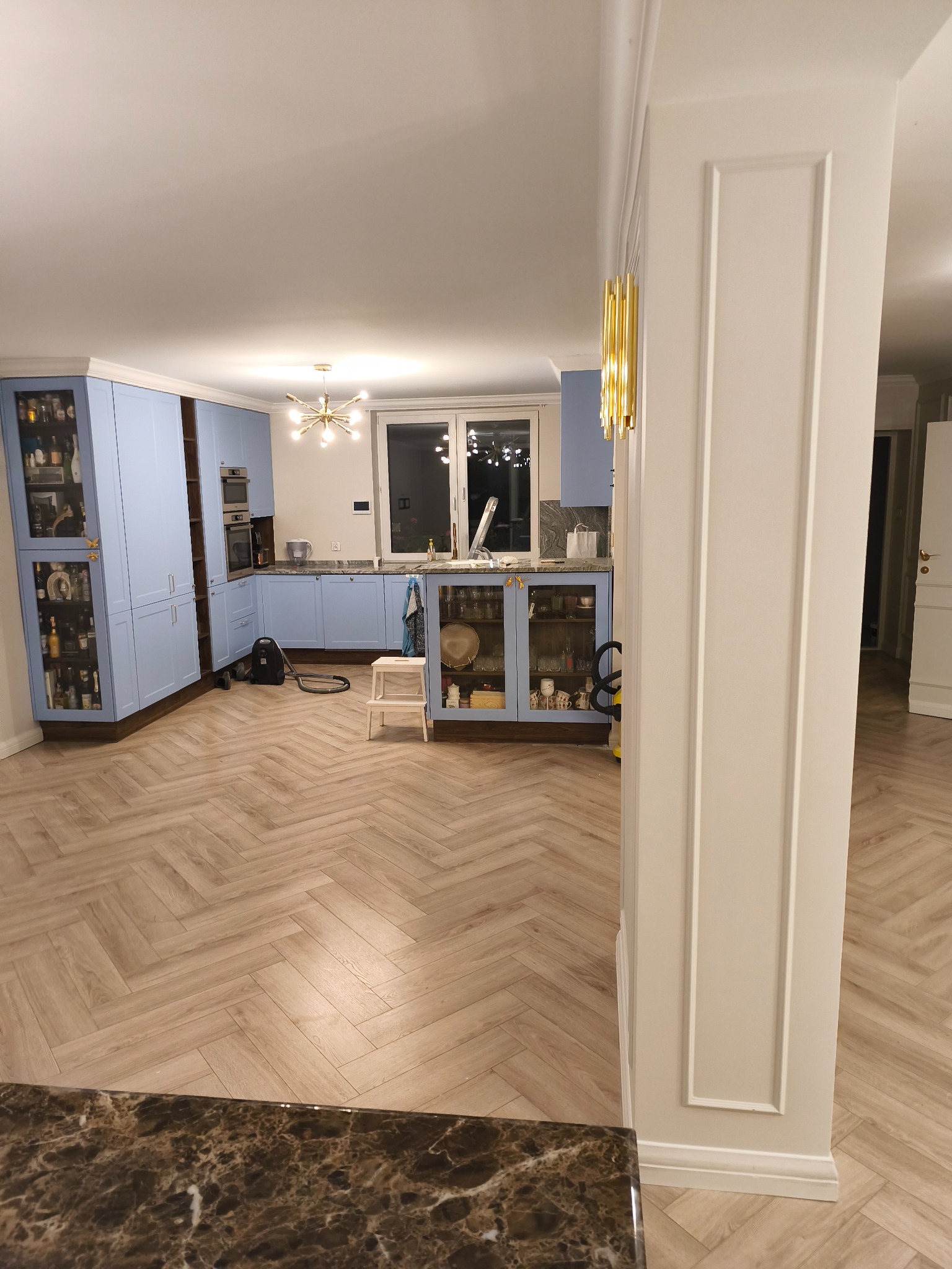 Jasna kuchnia w błękitnych odcieniach, z parkietem w jodełkę i złotymi akcentami oświetleniowymi. Widok z perspektywy blatu, z kolumną dekoracyjną po prawej.