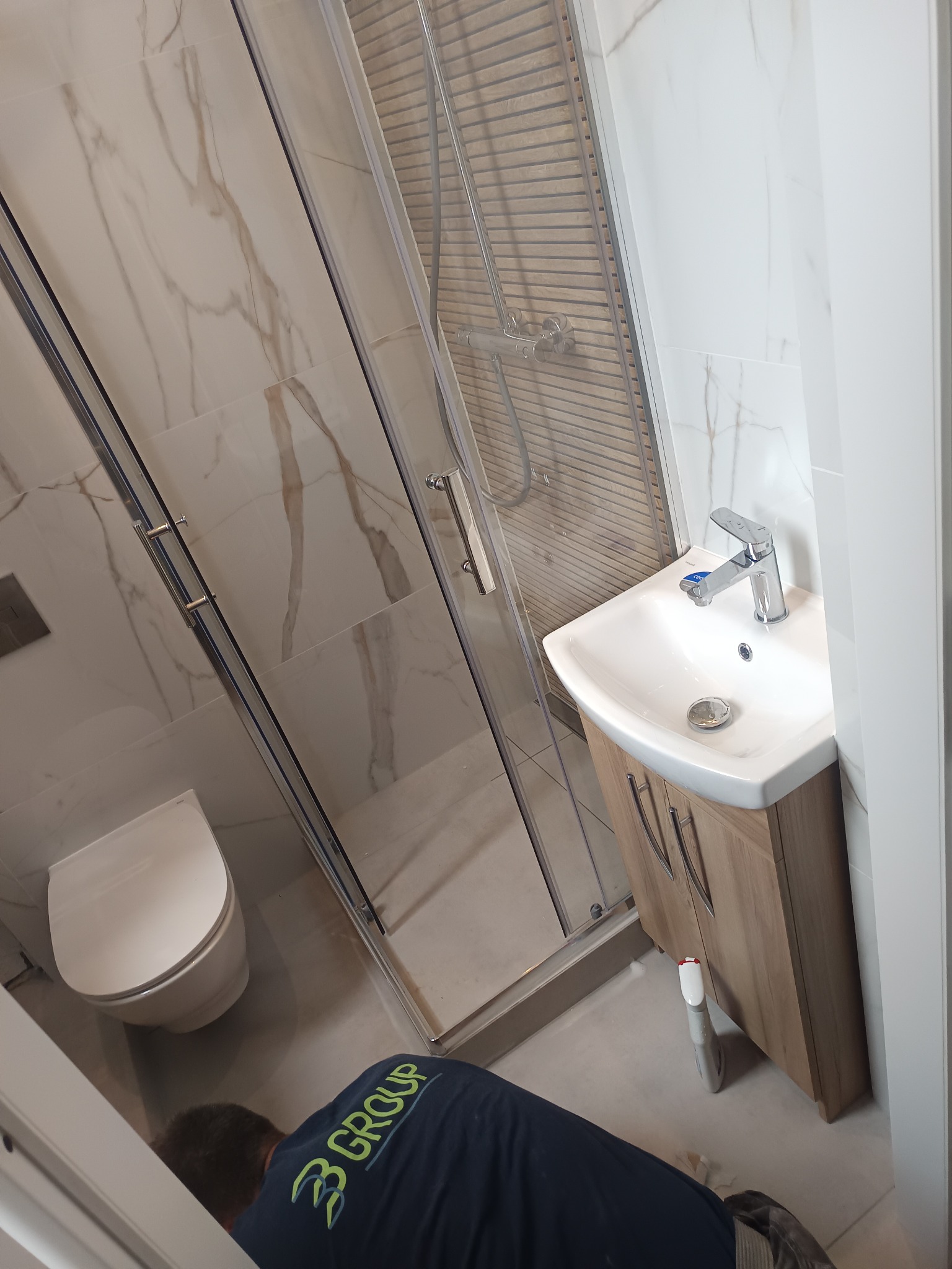 Wykończona łazienka z kabiną prysznicową, umywalką z szafką i toaletą. Białe marmurowe płytki na ścianach. Widoczny fragment osoby w koszulce z logo firmy.