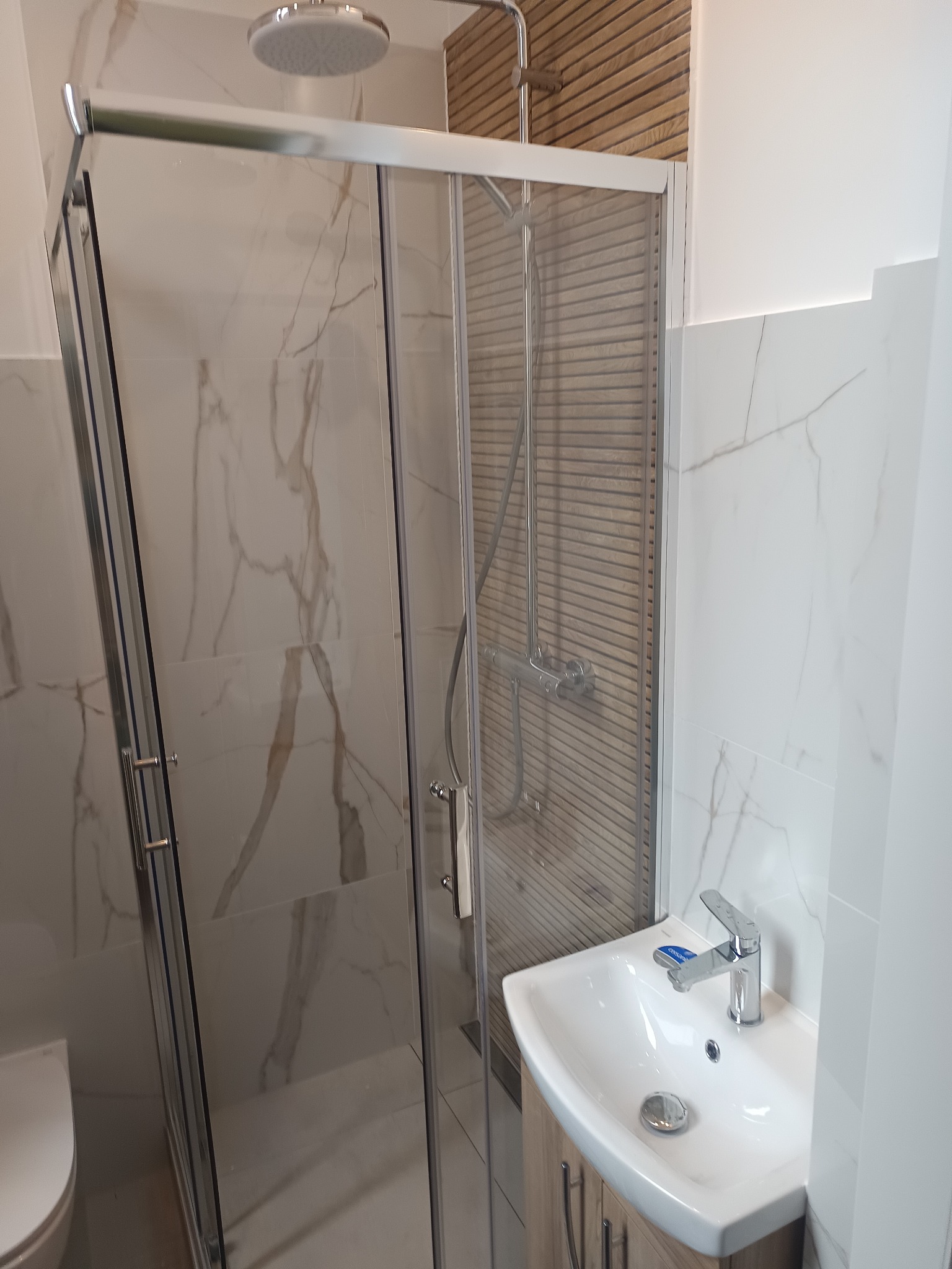 Wykończona łazienka z kabiną prysznicową, umywalką z szafką i marmurowymi płytkami. Widoczny panel dekoracyjny z drewnianych listewek. Nowoczesny design.