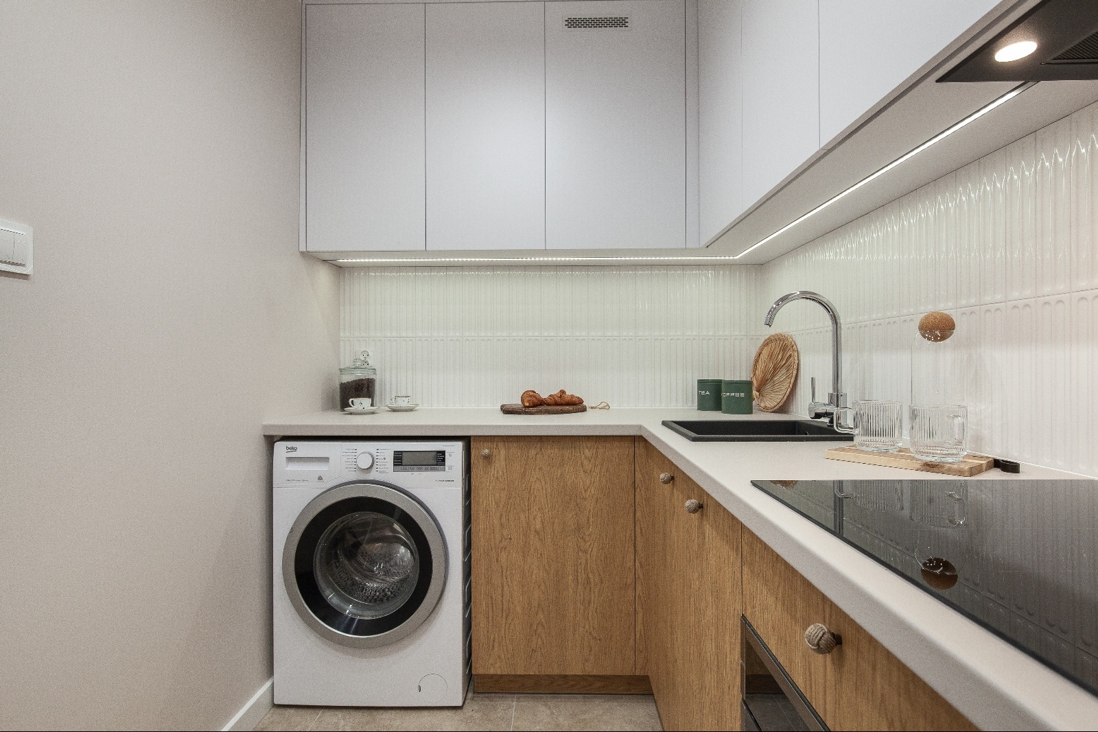 Nowoczesna kuchnia z pralką w zabudowie, drewniane fronty szafek, białe blaty i górne szafki, oświetlenie LED pod szafkami, minimalistyczny design, płytki z subtelnym wzorem.