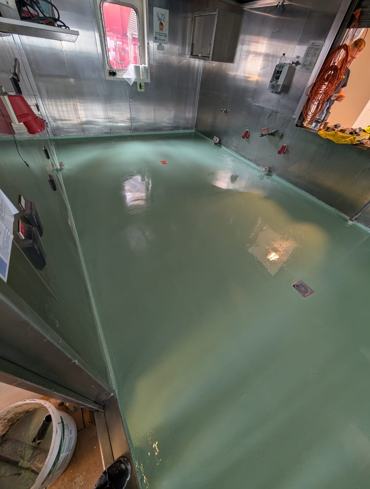 Świeżo pomalowana, gładka, zielona podłoga w pomieszczeniu o metalowych ścianach. Widoczne narzędzia i wiadro z farbą. Oświetlenie odbija się od mokrej powierzchni.