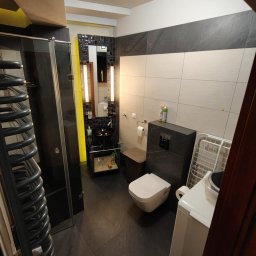 Homedetailingswarzedz - Nowoczesna łazienka z czarnymi płytkami podłogowymi, kabiną prysznicową i umywalką na żółtym stelażu. Widoczna pralka i grzejnik drabinkowy.