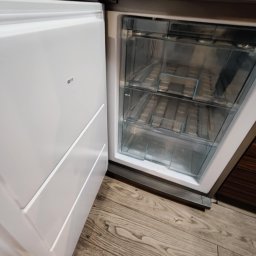 Homedetailingswarzedz - Otwarta, pusta zamrażarka z trzema przezroczystymi szufladami, widoczna na tle drewnianej podłogi i fragmentu zabudowy kuchennej. Minimalistyczny design.