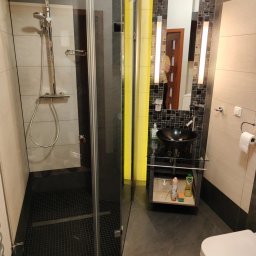 Homedetailingswarzedz - Nowoczesna łazienka z kabiną prysznicową typu walk-in, czarną umywalką na szklanym blacie i mozaiką nad lustrem. Widoczne środki czystości pod umywalką.