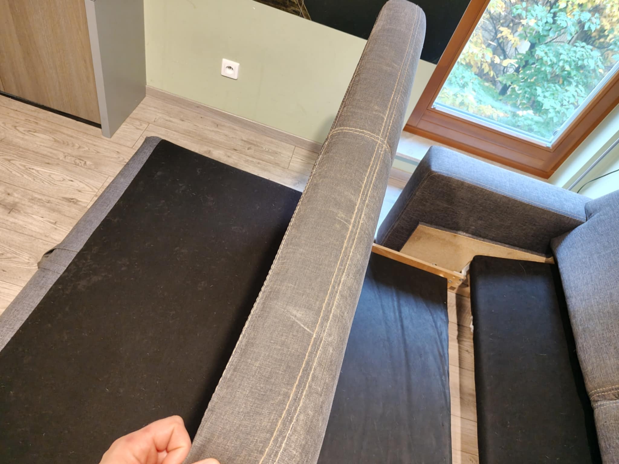 Szary narożnik z podniesionym siedziskiem, ukazujący mechanizm i wnętrze. Widoczne ślady użytkowania na tapicerce i konstrukcji. Ujęcie z góry, z oknem w tle.