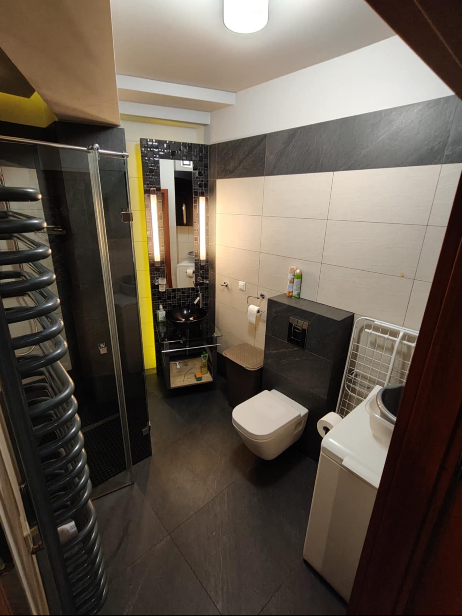 Nowoczesna łazienka z czarnymi płytkami podłogowymi, kabiną prysznicową i umywalką na żółtym stelażu. Widoczna pralka i grzejnik drabinkowy.