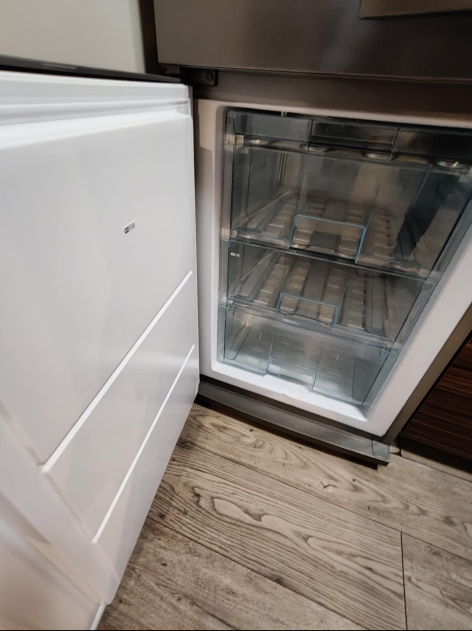 Otwarta, pusta zamrażarka z trzema przezroczystymi szufladami, widoczna na tle drewnianej podłogi i fragmentu zabudowy kuchennej. Minimalistyczny design.