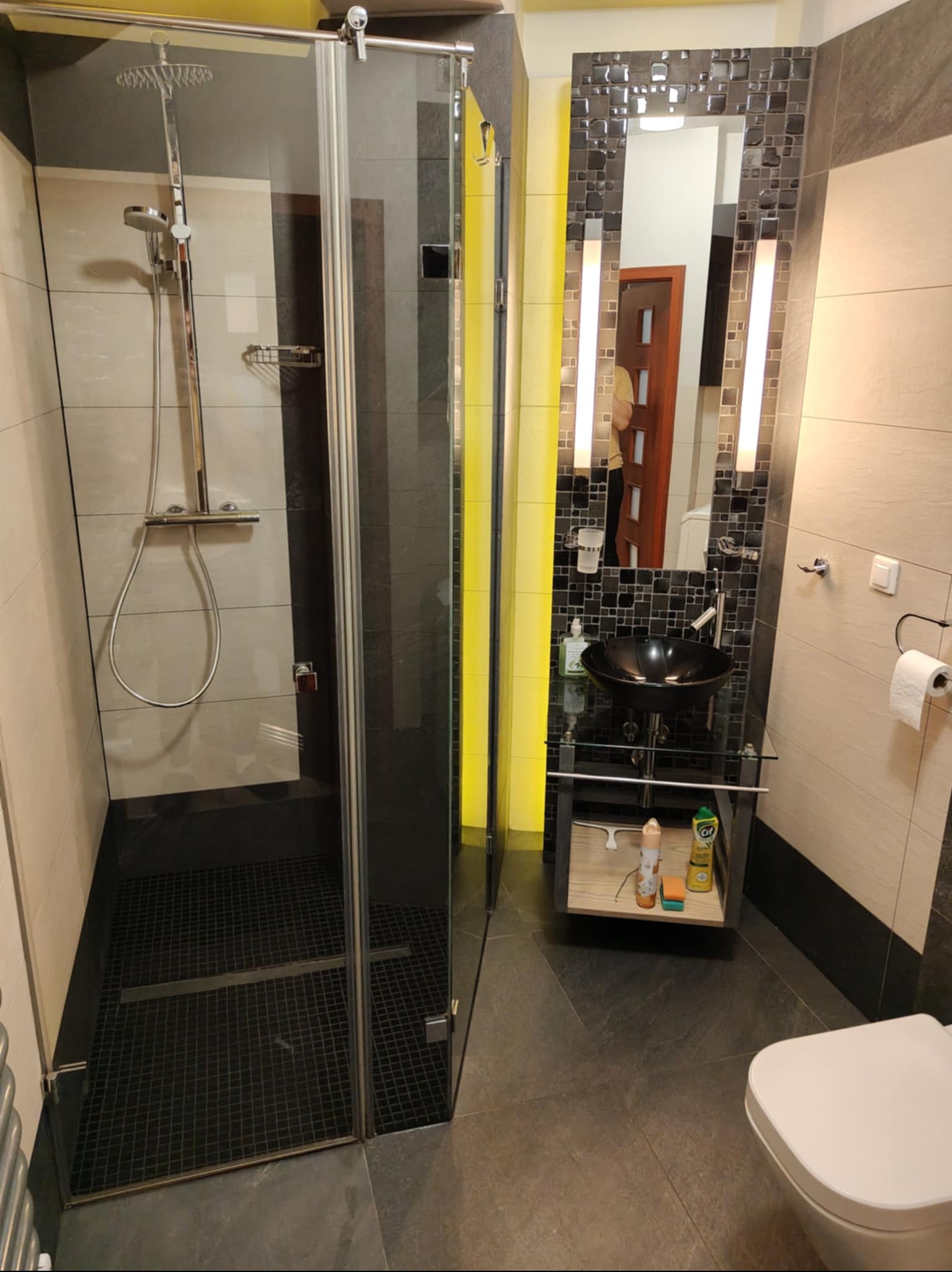 Nowoczesna łazienka z kabiną prysznicową typu walk-in, czarną umywalką na szklanym blacie i mozaiką nad lustrem. Widoczne środki czystości pod umywalką.