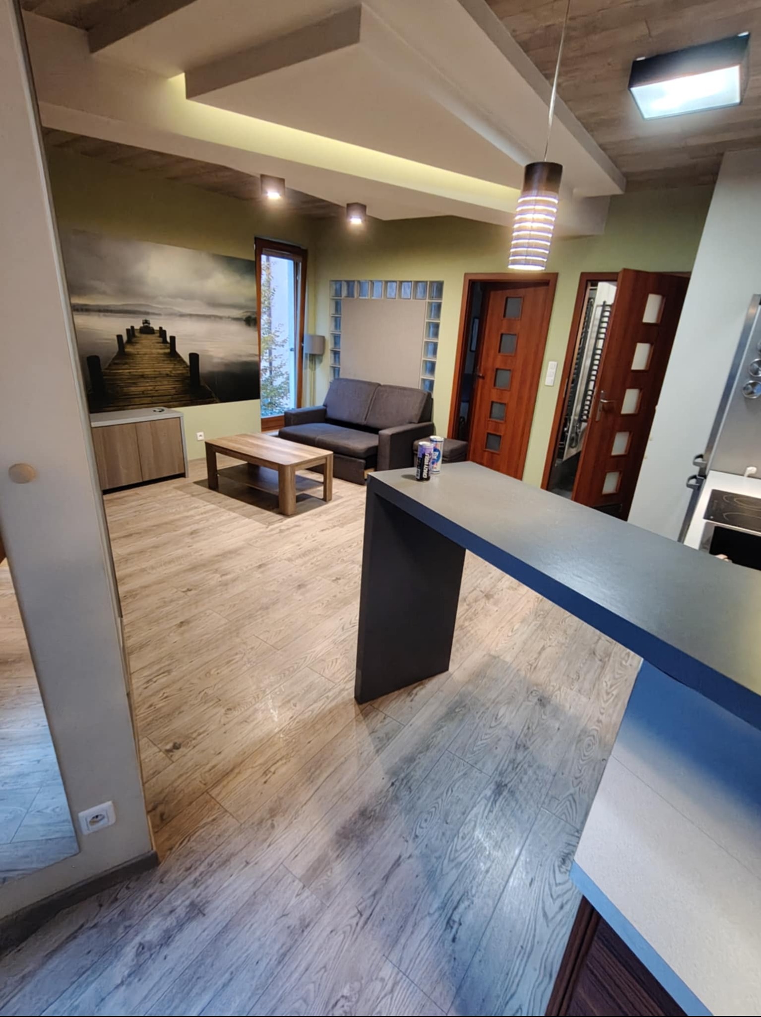 Jasne wnętrze salonu z drewnianą podłogą, szarą sofą i obrazem mola na ścianie. Widoczny fragment blatu kuchennego i lampa wisząca. Dwa napoje na blacie.