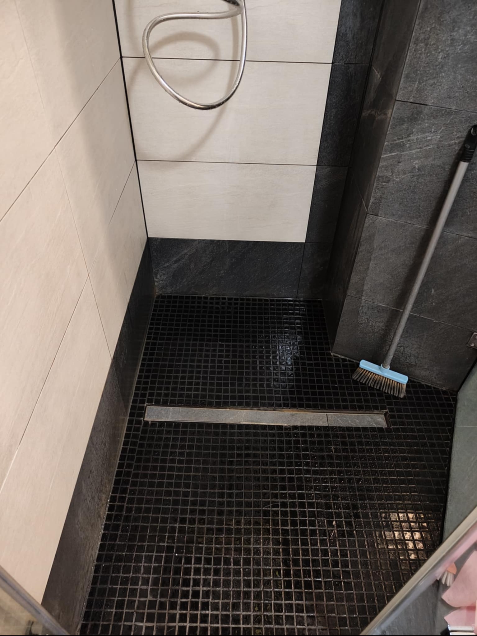 Umyta kabina prysznicowa z czarną mozaiką podłogową, odpływem liniowym i mopem opartym o ścianę. Widoczne płytki w odcieniach bieli i szarości.
