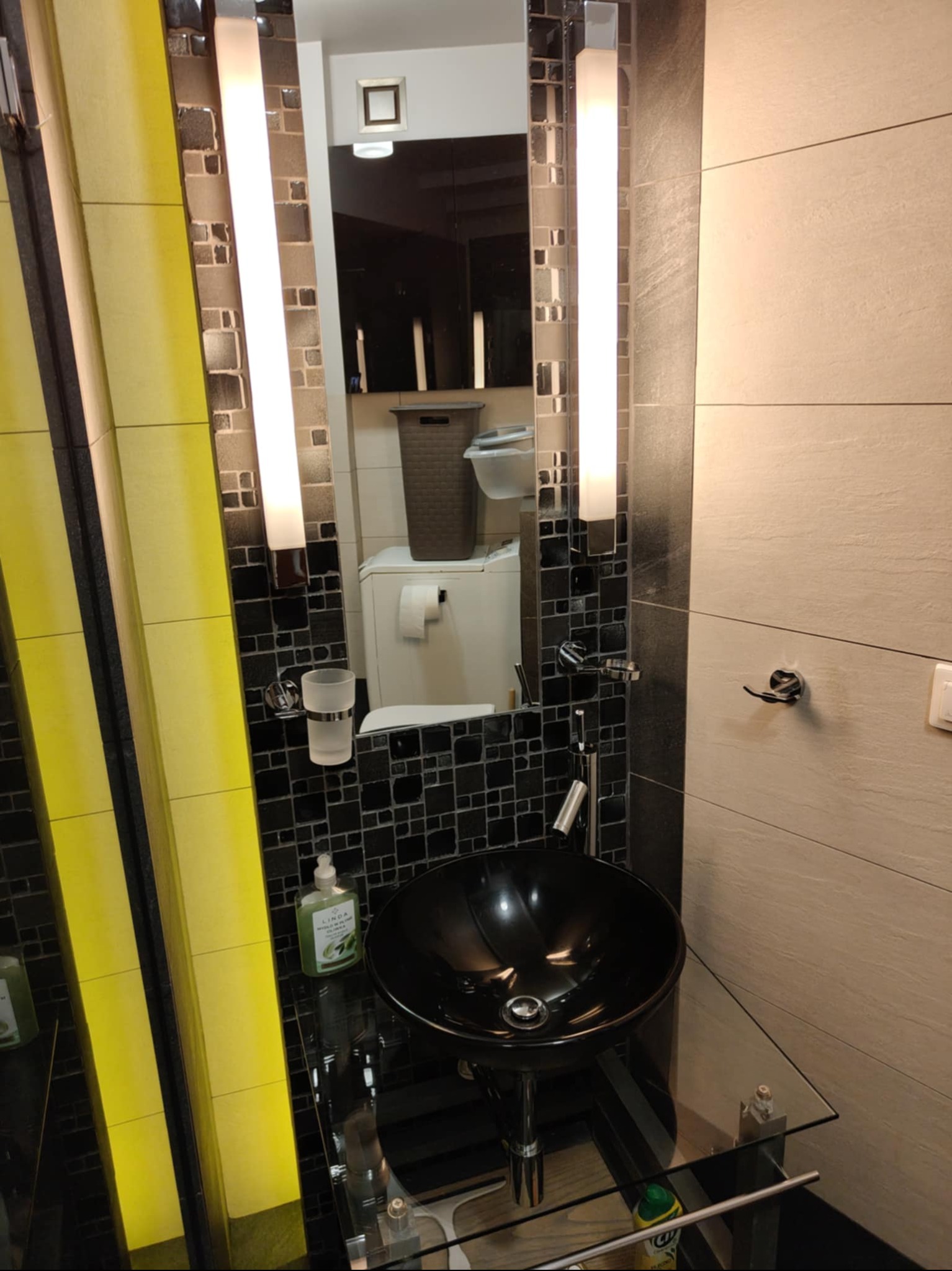 Nowoczesna łazienka z czarną umywalką na szklanym blacie, oświetlona pionowymi lampami. W tle widoczne płytki mozaikowe i pralka.