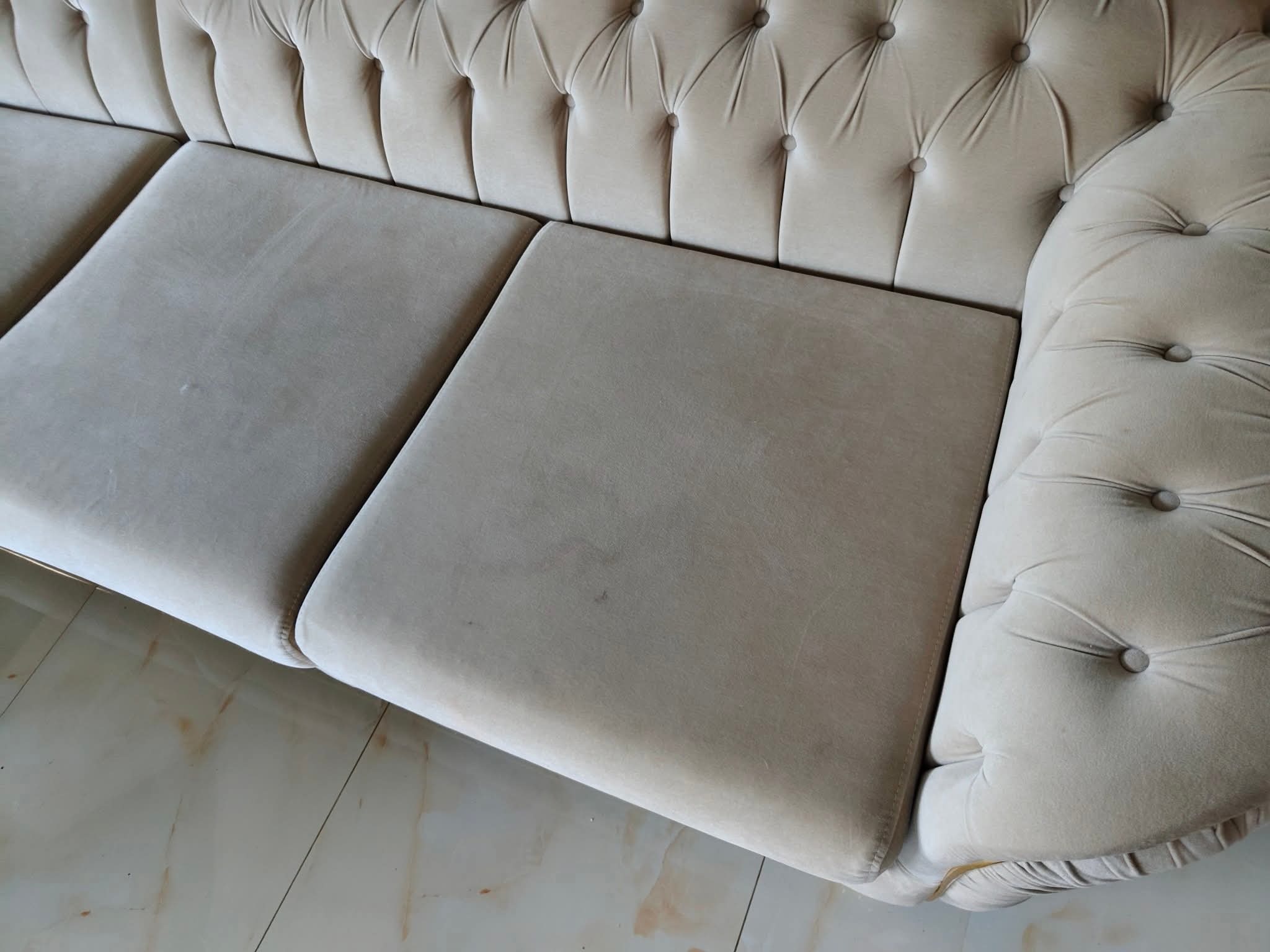 Beżowa sofa z pikowanym oparciem i podłokietnikiem, widoczna z góry, na marmurowej podłodze. Widoczne lekkie zabrudzenia na siedzisku.