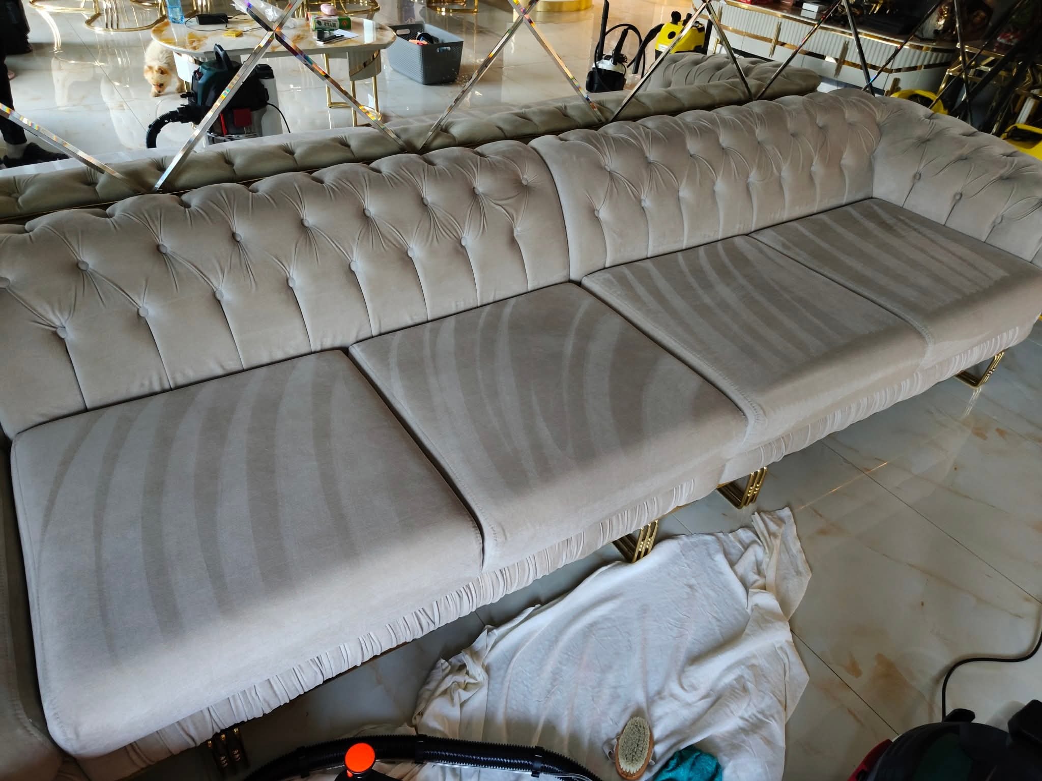 Beżowa, pikowana sofa po praniu tapicerki, widoczne ślady czyszczenia na siedziskach. W tle lustrzana ściana i elementy wyposażenia wnętrza. Na podłodze akcesoria do prania.