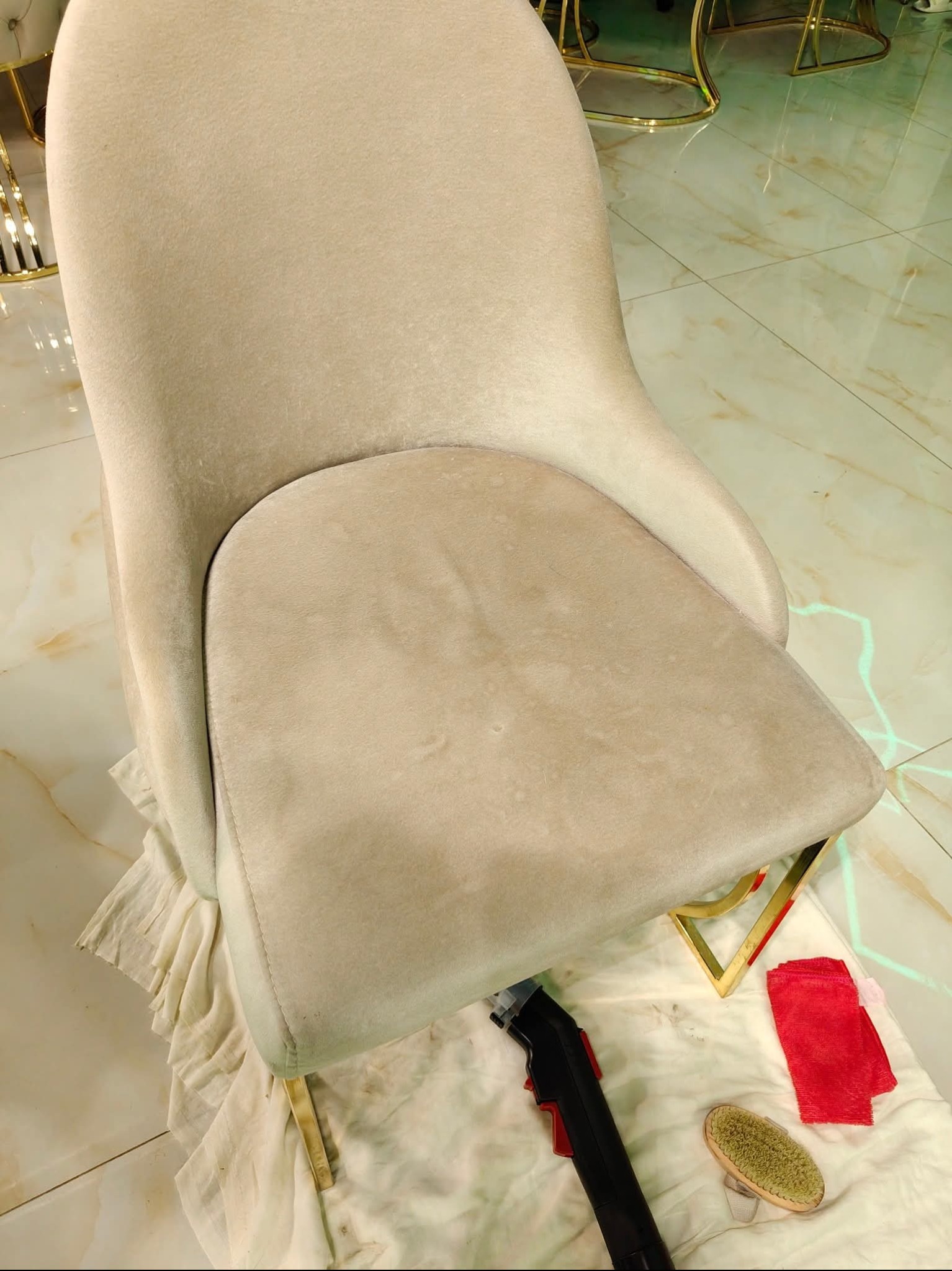 Kremowe krzesło tapicerowane z widocznymi plamami na siedzisku, przygotowane do czyszczenia. Obok leżą narzędzia: odkurzacz piorący, szczotka i czerwona ściereczka.