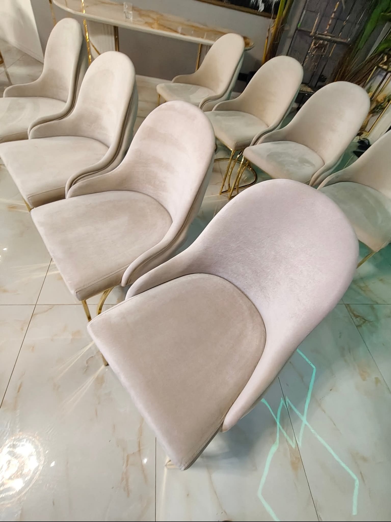 Rząd beżowych krzeseł tapicerowanych z metalowymi nogami w złotym kolorze, ustawionych na błyszczącej podłodze z marmuru. Krzesła o zaokrąglonych oparciach.
