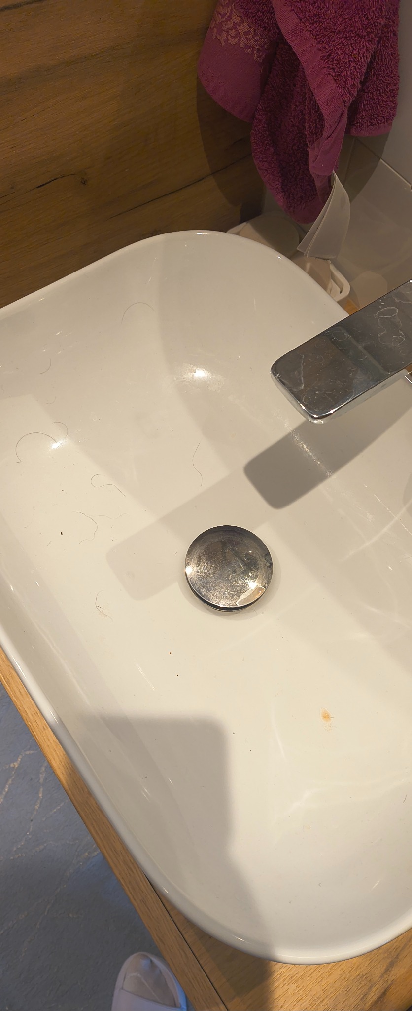 Biała umywalka z widocznymi włosami i zabrudzeniami, chromowany kran i odpływ. W tle drewniana ściana i fioletowy ręcznik. Ujęcie z góry.