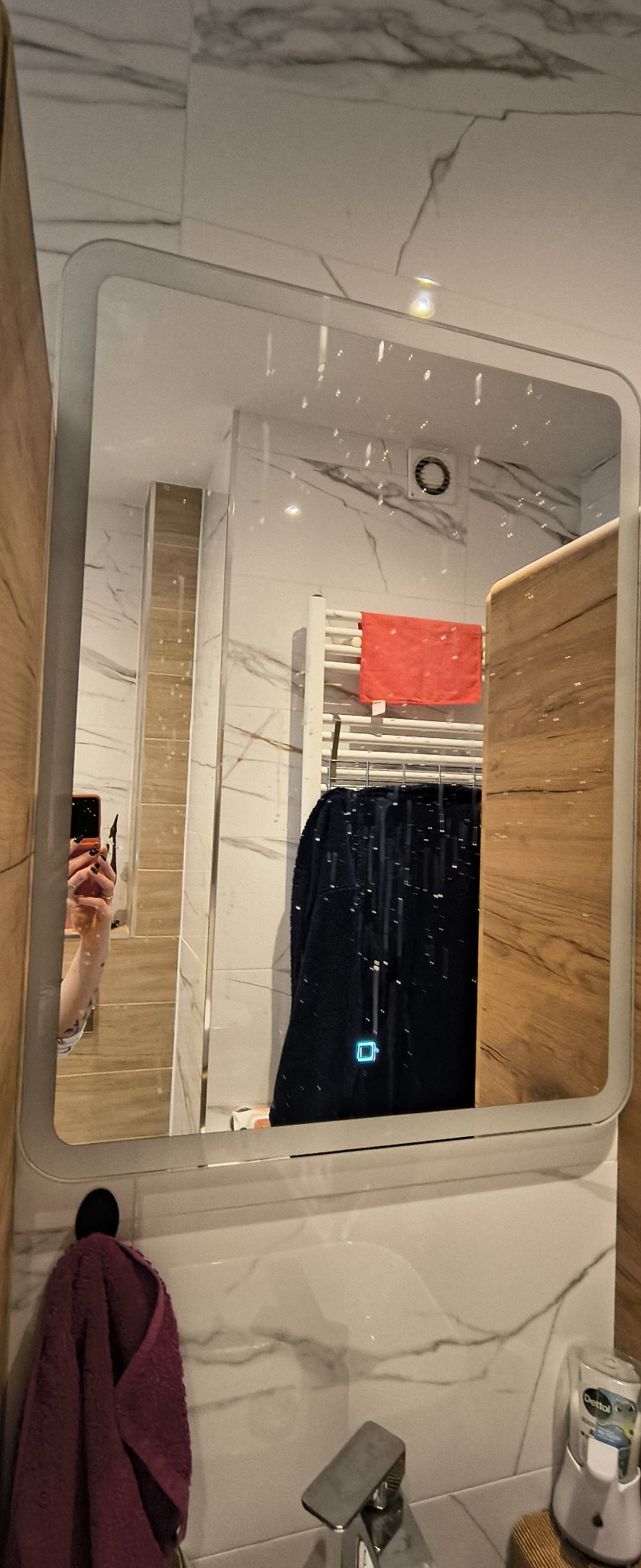 Nowoczesna łazienka z marmurowymi płytkami, widoczna w lustrze z podświetleniem LED. Na grzejniku suszą się ręczniki, a obok umywalka z dozownikiem mydła.