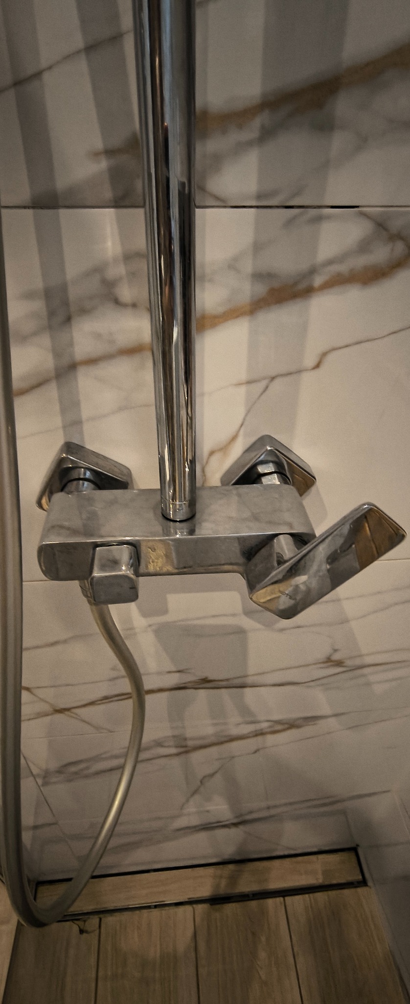 Błyszcząca, chromowana bateria prysznicowa z widocznymi śladami po czyszczeniu, na tle marmurowej ściany i drewnopodobnej podłogi z odpływem liniowym.