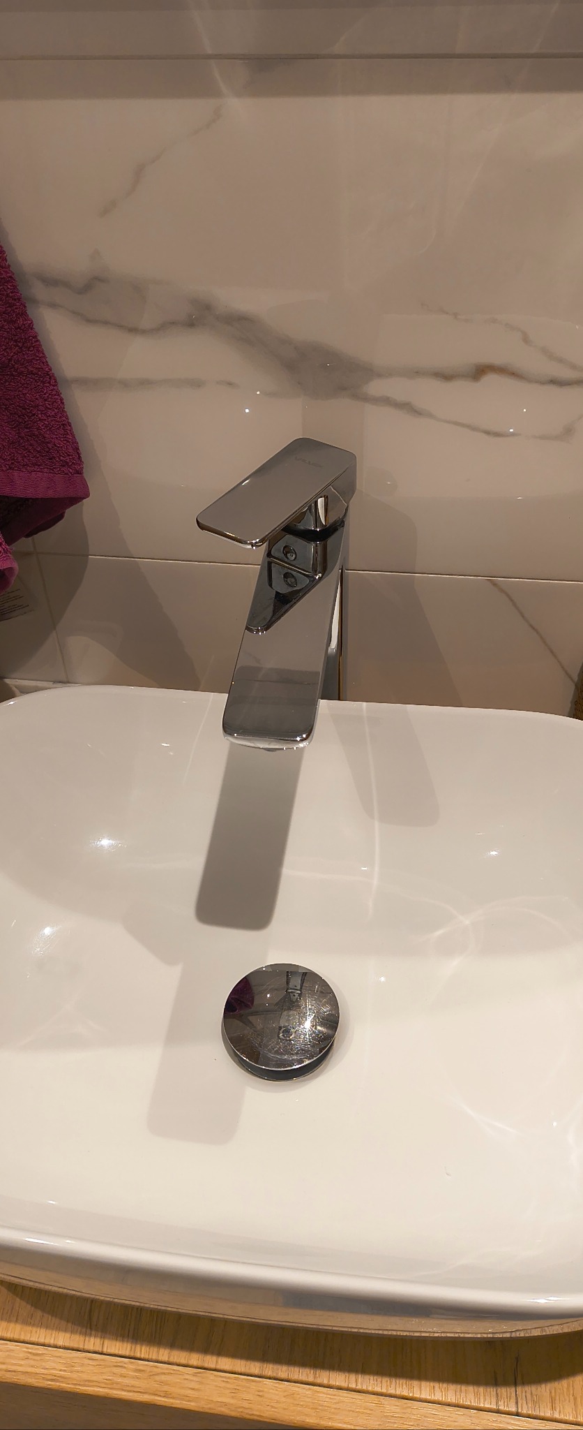 Błyszcząca, chromowana bateria umywalkowa na białej, ceramicznej umywalce z odpływem. W tle marmurowe płytki i fragment fioletowego ręcznika.