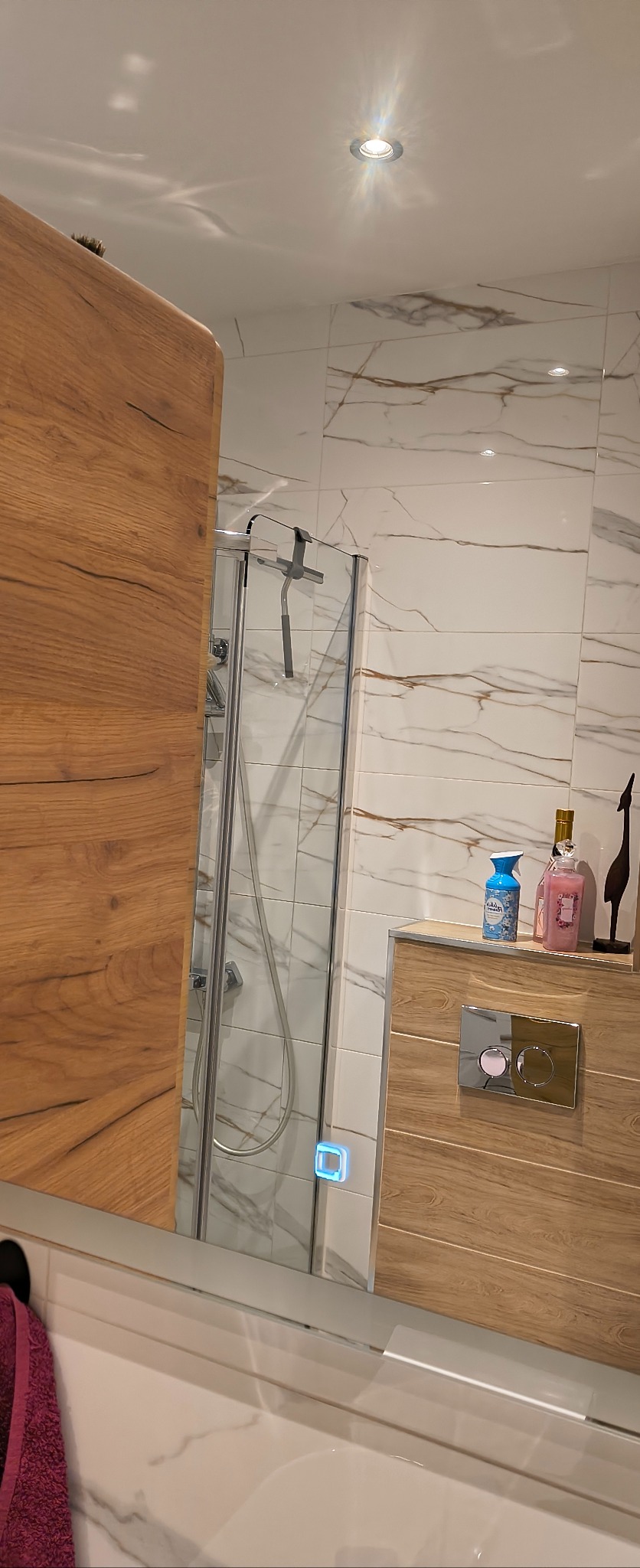 Błyszcząca kabina prysznicowa za drewnianym blatem z dozownikami i dekoracją, odbijająca się w lustrze. Białe płytki z brązowymi żyłkami w tle.