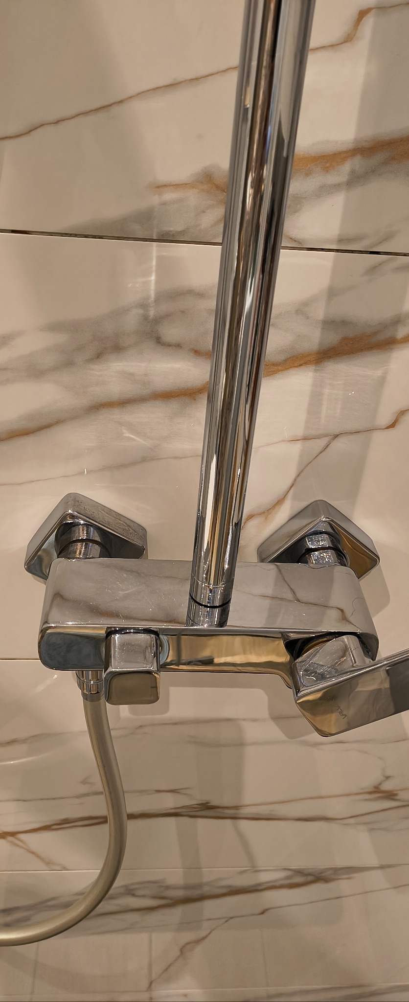 Błyszcząca, chromowana bateria prysznicowa z wężem, zamontowana na marmurowej ścianie. Widok z dołu, ukazujący detale armatury i wzór kamienia.