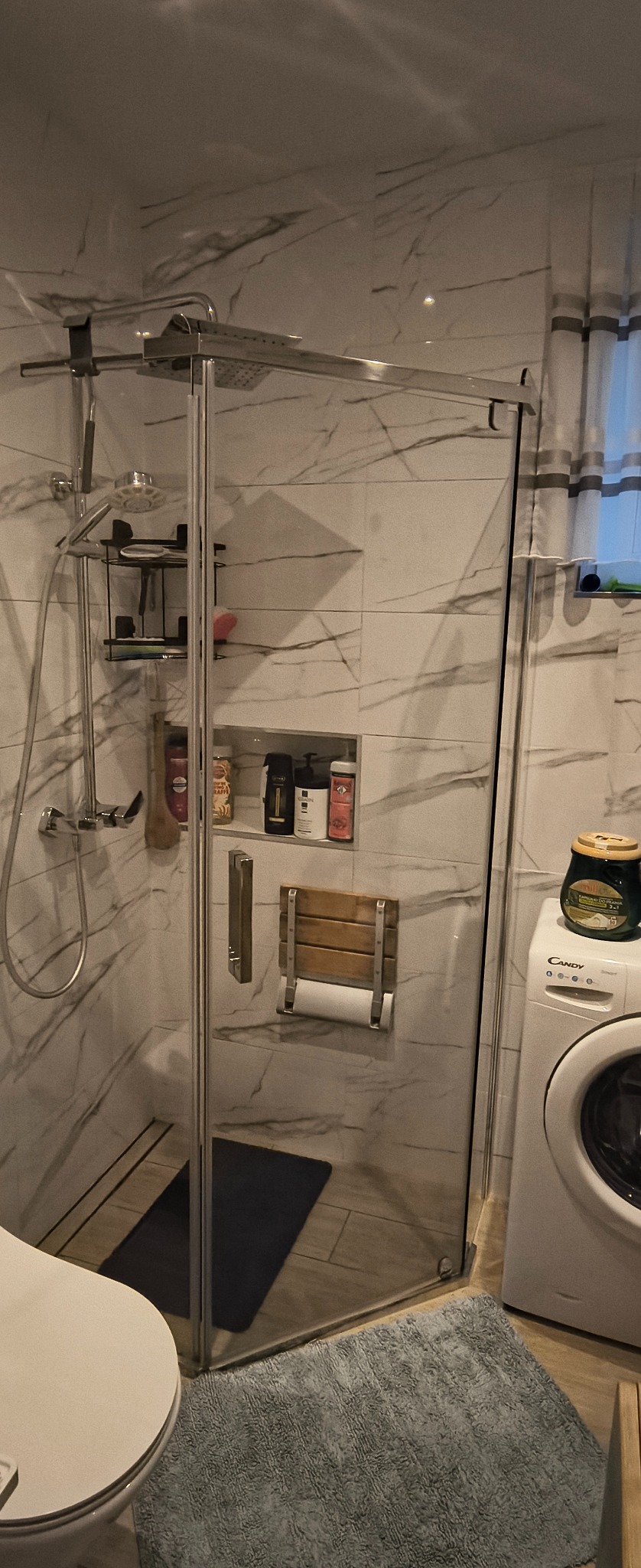 Nowoczesna kabina prysznicowa z marmurowymi płytkami, drewnianym siedziskiem i wnęką na kosmetyki. Obok pralka i dywanik. Widok na łazienkę po remoncie.