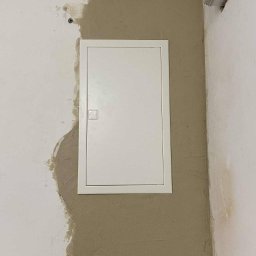 MMC-elektryka Michał Czyż - Biała skrzynka elektryczna w ścianie po szpachlowaniu, widoczne ślady świeżego gładzi. Ujęcie z bliska, neutralne oświetlenie.