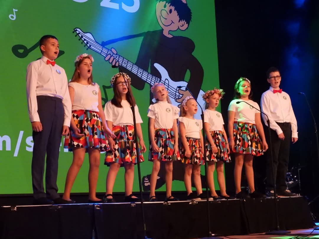 Występ chóru dziecięcego na scenie z gitarą w tle. Dzieci ubrane w białe koszulki i kolorowe spódniczki, śpiewają do mikrofonów. Koncert.