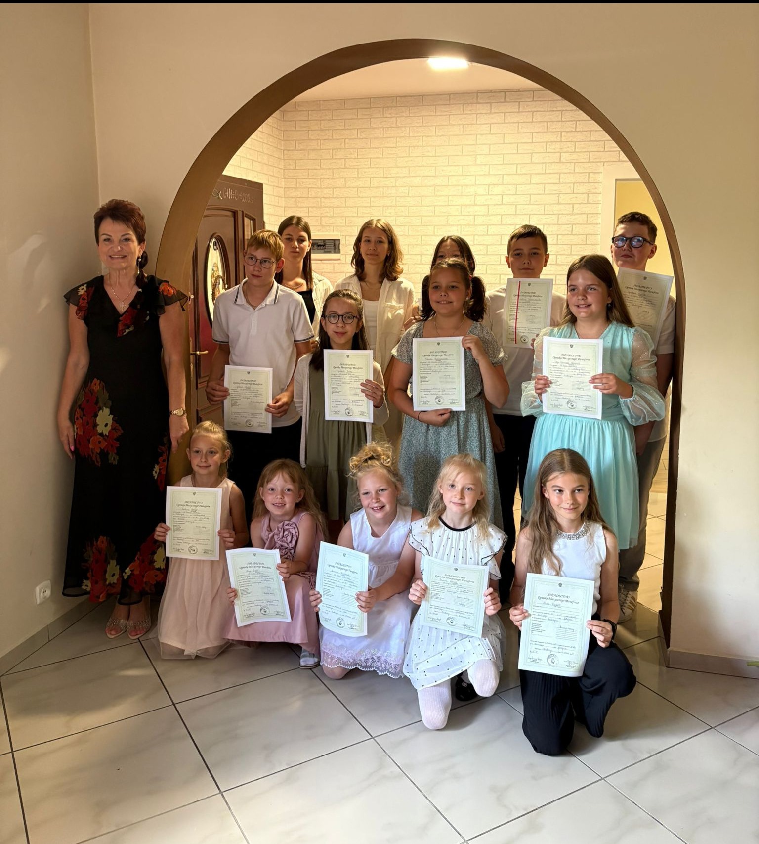 Grupa dzieci i dorosła kobieta, stojący i siedzący, prezentują certyfikaty ukończenia kursu, prawdopodobnie muzycznego, na tle białej ściany z cegieł i łukowatego przejścia.
