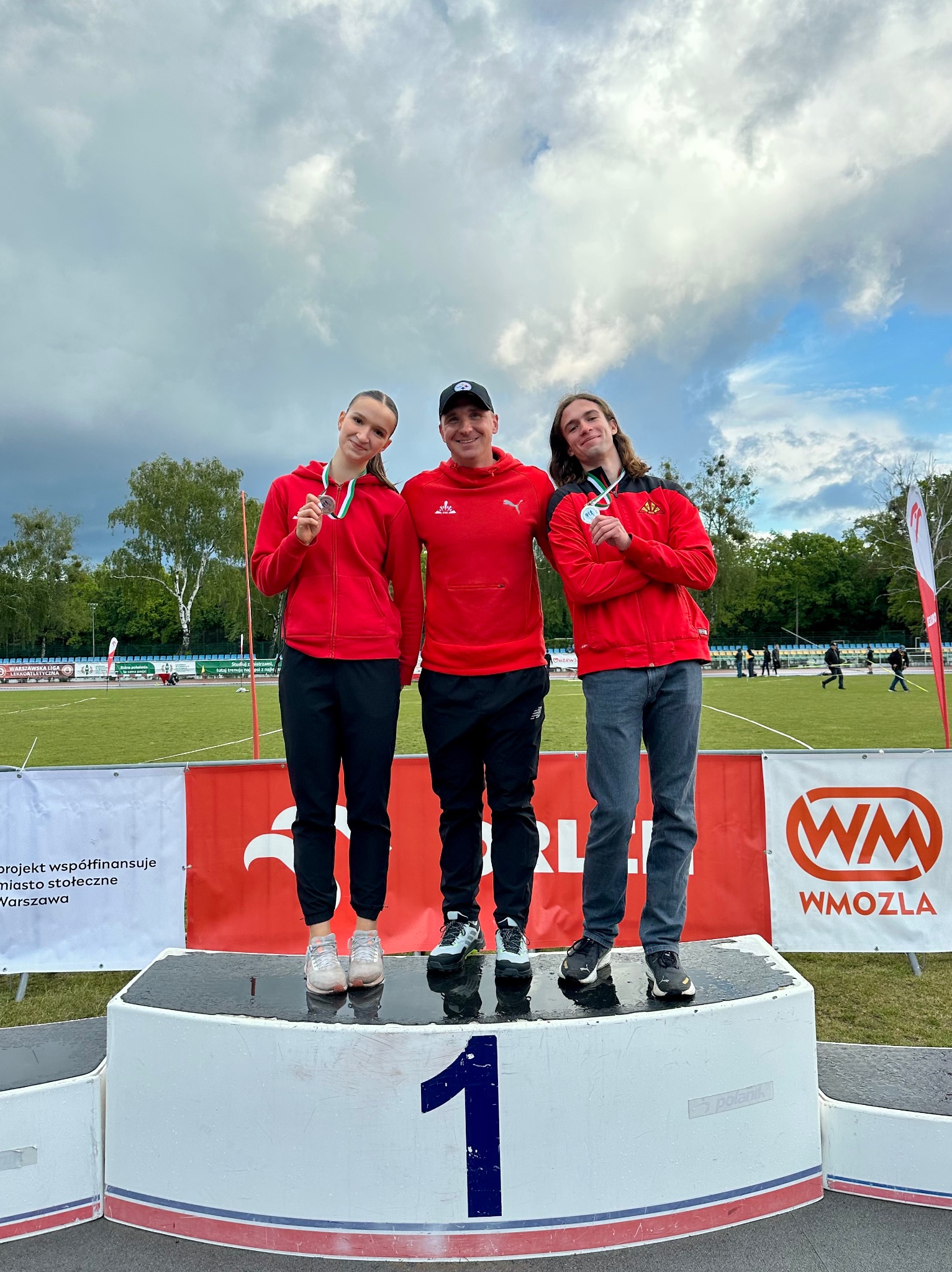 Trójka sportowców na podium z medalami, w czerwonych bluzach, na tle stadionu lekkoatletycznego i pochmurnego nieba. Trener w środku.
