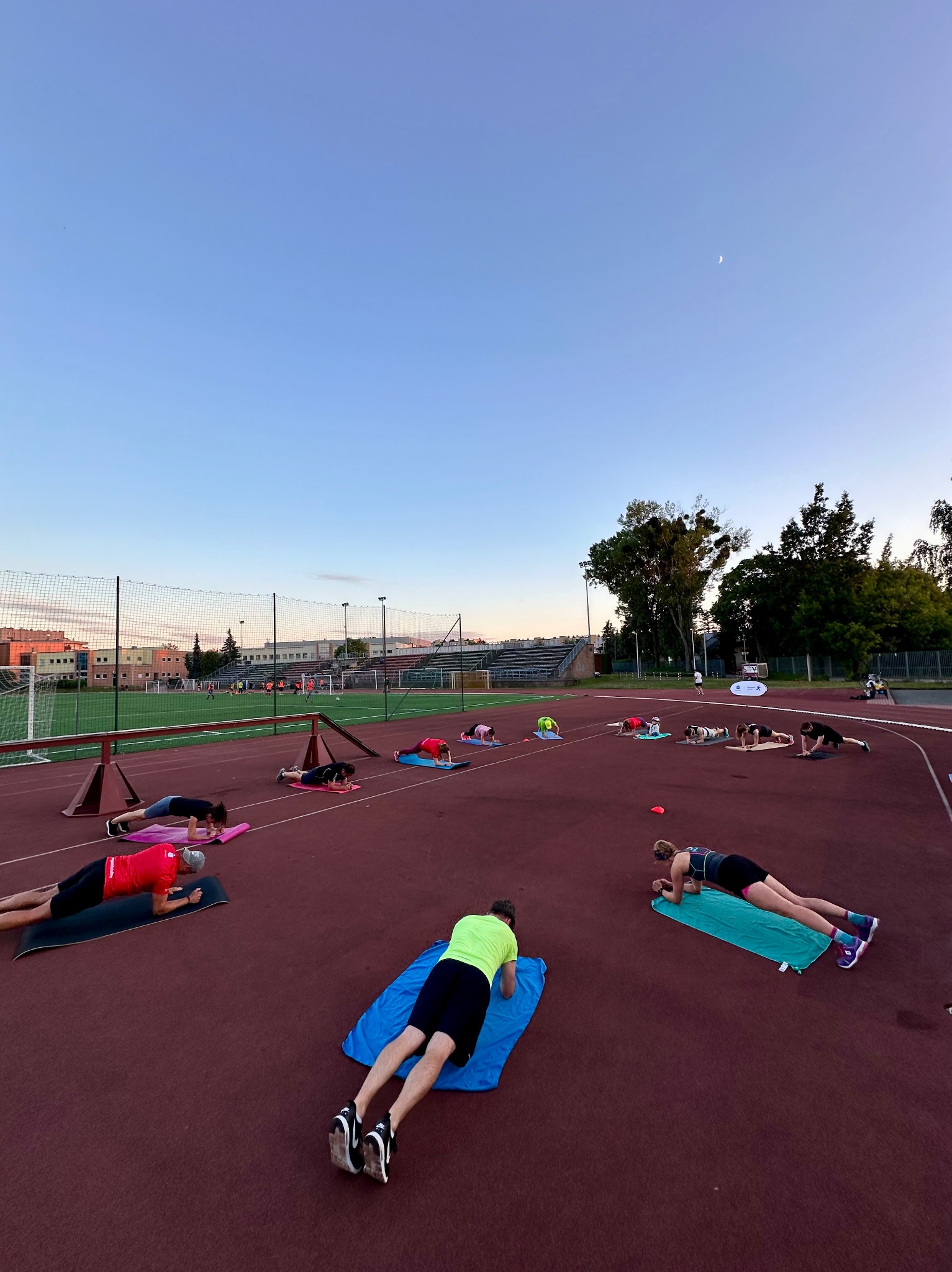 Grupowy trening na stadionie: osoby w pozycji plank na matach, w tle bieżnia, trybuny i budynki. Błękitne niebo z widocznym księżycem w tle.