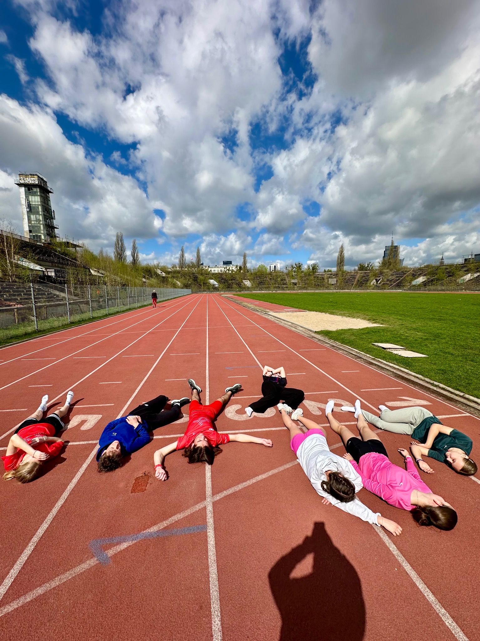 Grupa osób leży na bieżni stadionu, relaksując się po treningu. W tle widoczna wieża stadionu i błękitne niebo z chmurami. Kąt ujęcia z góry podkreśla perspektywę bieżni.
