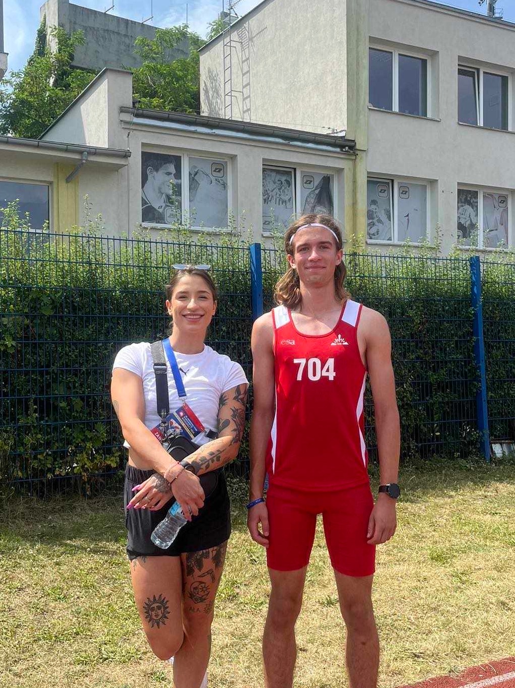Uśmiechnięta kobieta z tatuażami i mężczyzna w stroju sportowym z numerem 704 stoją na tle budynku z plakatami w oknach i zieleni. Trening biegowy na świeżym powietrzu.