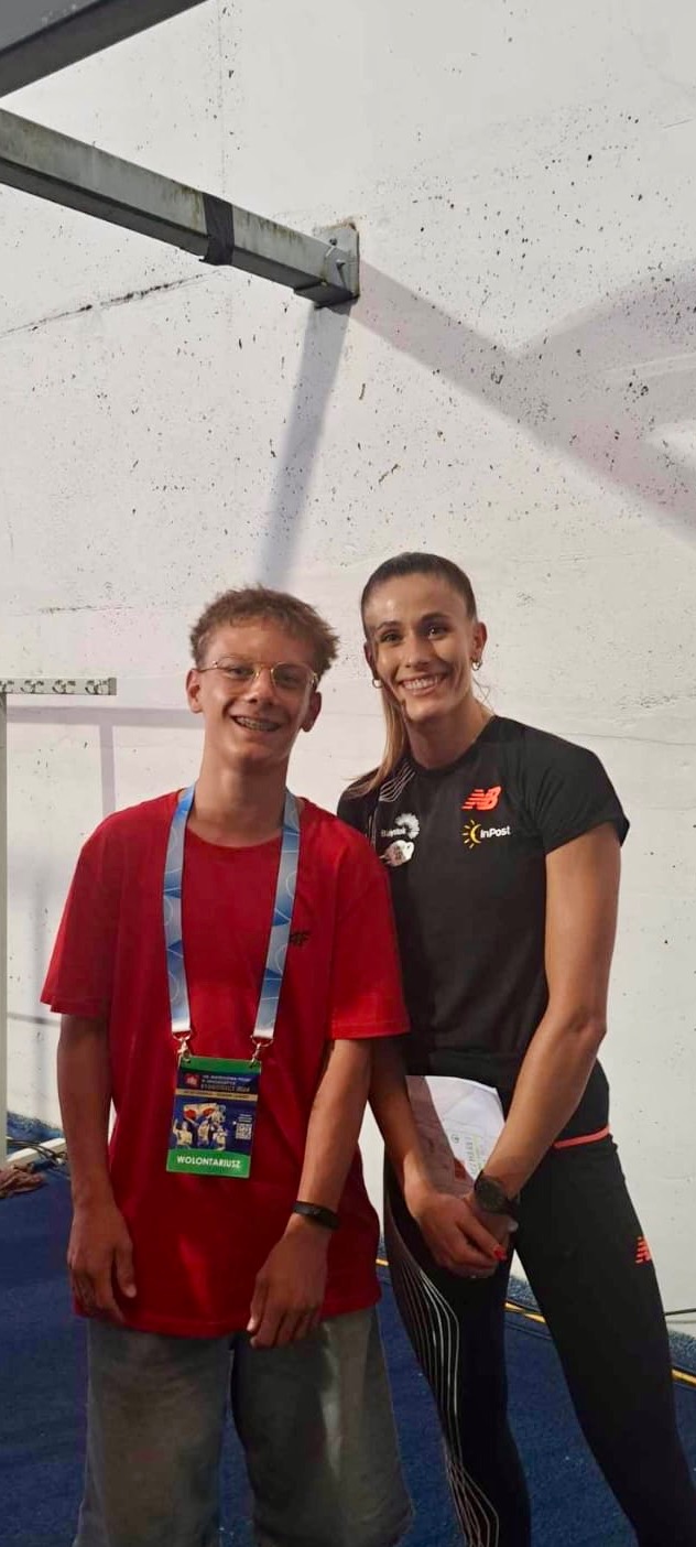 Uśmiechnięta lekkoatletka w czarnej koszulce z logo InPost i New Balance pozuje z młodym wolontariuszem w czerwonej koszulce z identyfikatorem na tle białej ściany.