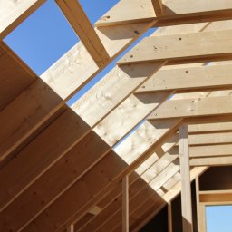 BUDUJE-GRUPA Spółka z ograniczoną odpowiedzialnością - Konstrukcja drewniana dachu w trakcie budowy, widok z dołu na jasne belki i fragment błękitnego nieba. Elementy złączone śrubami, widoczne detale drewna.